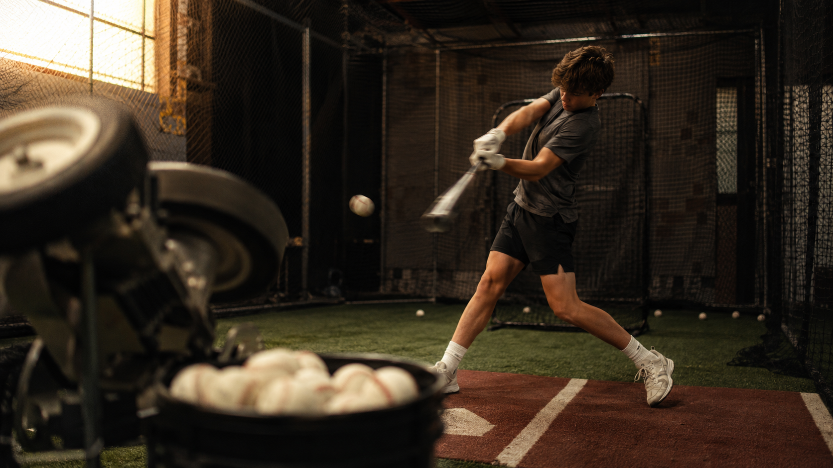 Hitter mid-swing in an indoor batting cage with a pitching machine and a bucket of Ventur3 baseballs in the foreground